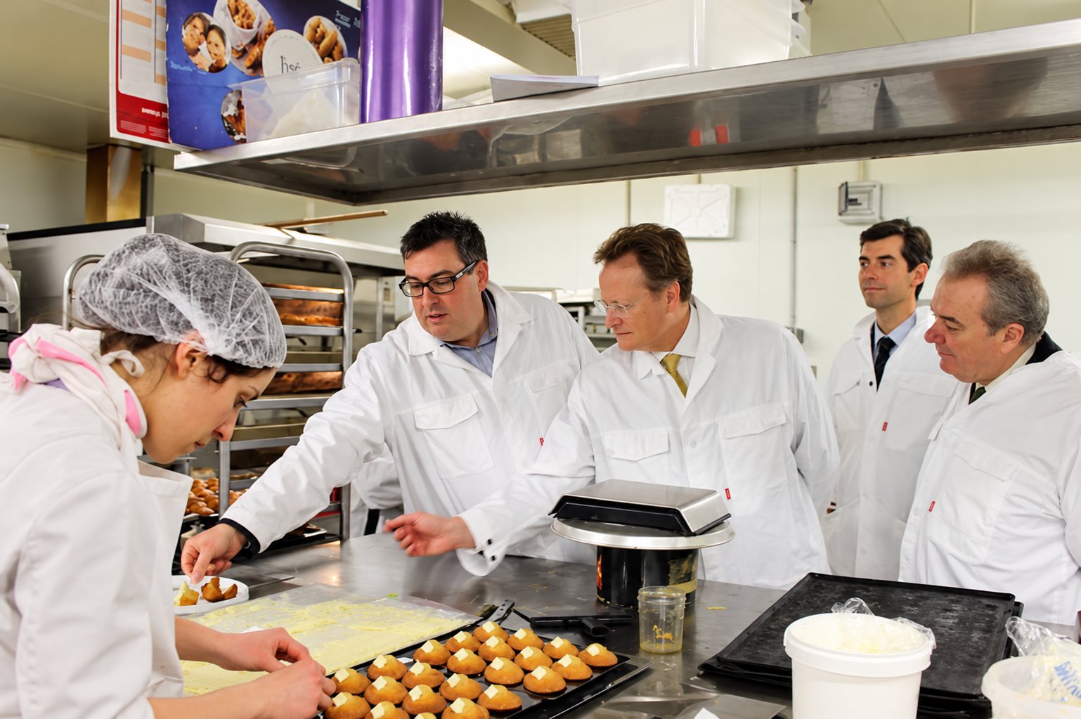 Dans un laboratoire de pâtisserie, une employée portant une charlotte prépare des petits gâteaux tandis que quatre hommes en blouse blanche l’observent.